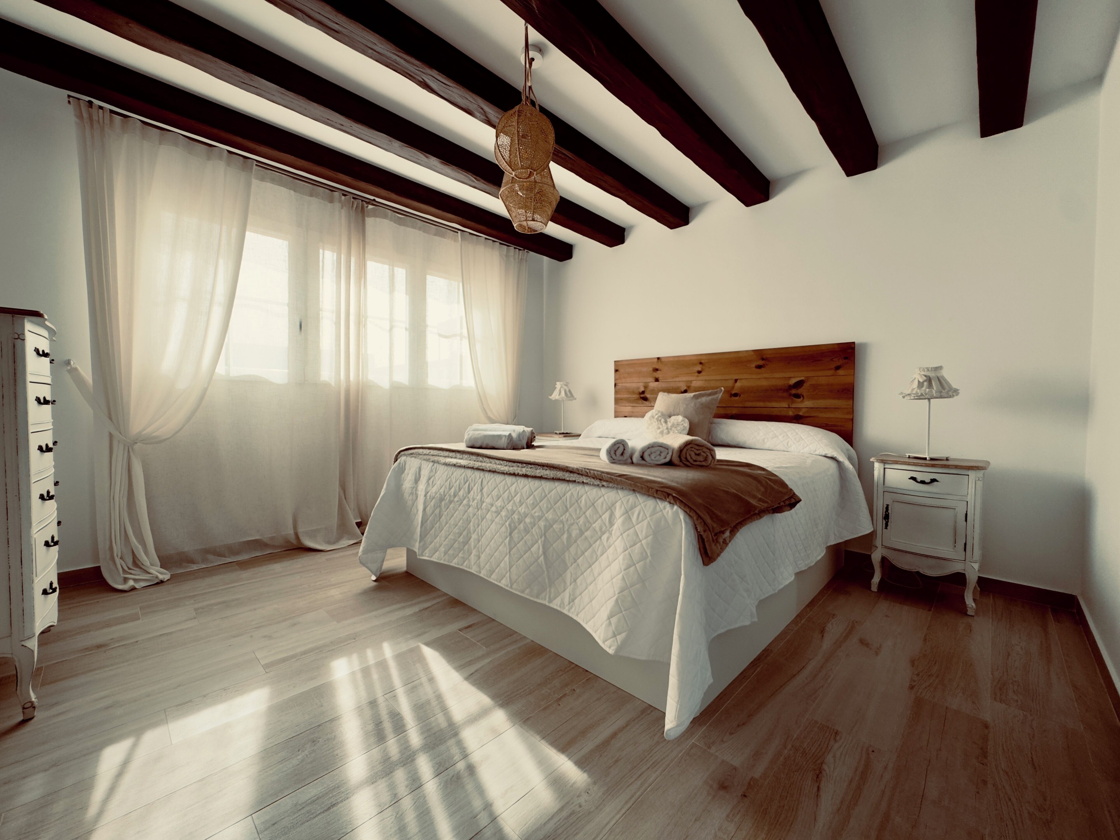 A bedroom with a wooden ceiling, a bed with a quilted bedspread, a nightstand with a lamp, and a window with sheer curtains.