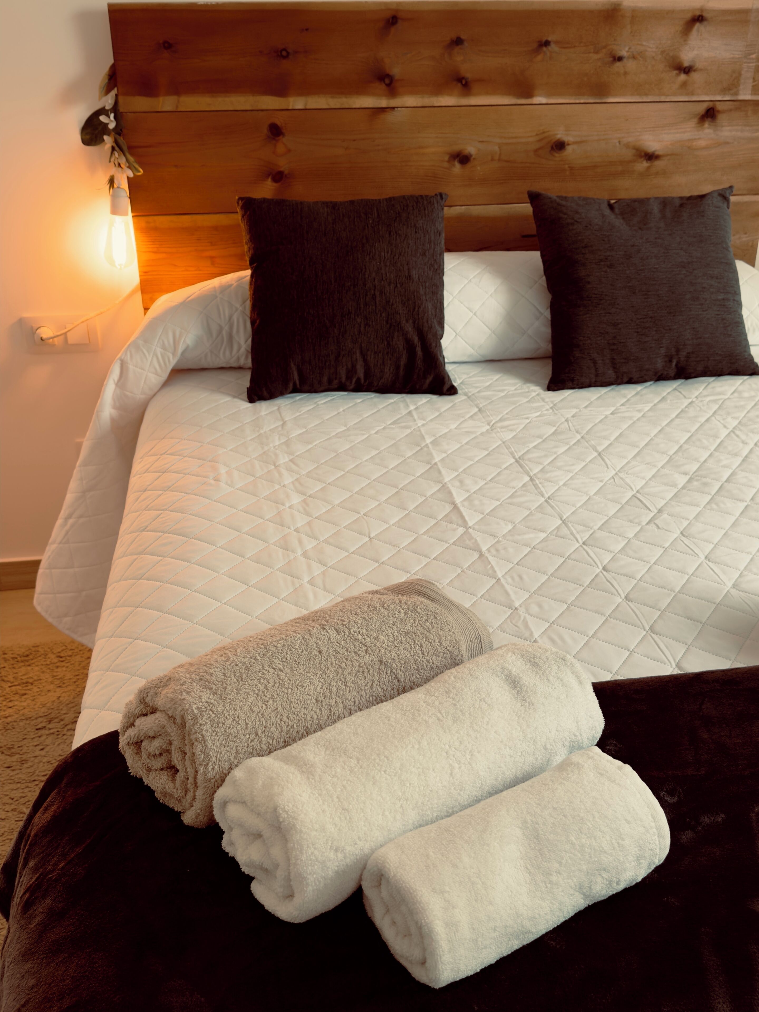 A bed with a quilted white duvet, two dark pillows, and a wooden headboard. There are two rolled towels on a dark ottoman in front of the bed.