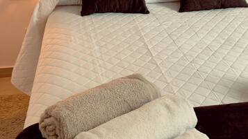 Una cama con edredón blanco acolchado, dos almohadas oscuras y un cabecero de madera. Hay dos toallas enrolladas sobre un otomano oscuro frente a la cama.