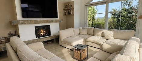 A living room with a sectional sofa, a fireplace, a flat-screen TV, and a large window with a view of greenery.