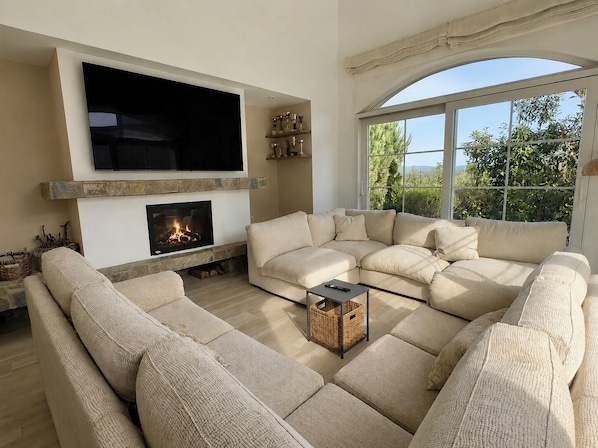 A living room with a sectional sofa, a fireplace, a flat-screen TV, and a large window with a view of greenery.