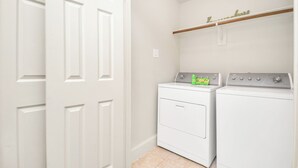 A laundry room with two white washers and dryers, a wooden shelf above the dryers, and a white door. - Afrotique HOU Getaway Pool!Galleria!Uptown (Houston)