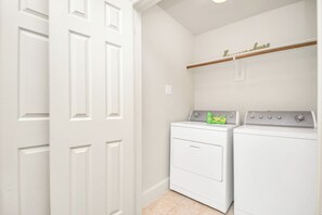 A laundry room with two white washers and dryers, a wooden shelf above the dryers, and a white door. - Afrotique HOU Getaway Pool!Galleria!Uptown (Houston)