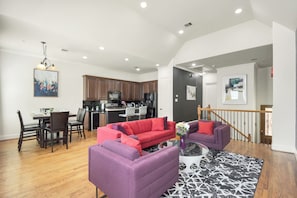 A modern living room with a red sofa, a purple sofa, and a coffee table. There is a dining area with a table and chairs. The kitchen is visible in the background with wooden cabinets and a black countertop. - Afrotique HOU Getaway Pool!Galleria!Uptown (Houston)