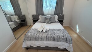 A modern bedroom with a large bed, bedside tables, a mirror, and a view of the outdoors through the window. - Your Ideal Stay Near Disney (Placentia)