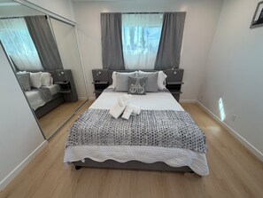 Ein modernes Schlafzimmer mit einem großen Bett, Nachttischen, einem Spiegel und Blick ins Freie durch das Fenster.