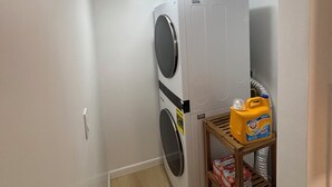 A laundry room with a stacked washer and dryer, a shelf with cleaning supplies, and a wooden floor. - Your Ideal Stay Near Disney (Placentia)
