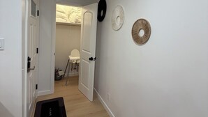 A hallway with a black mat, white walls, and a door leading to a room with a bed and a chair. - Your Ideal Stay Near Disney (Placentia)