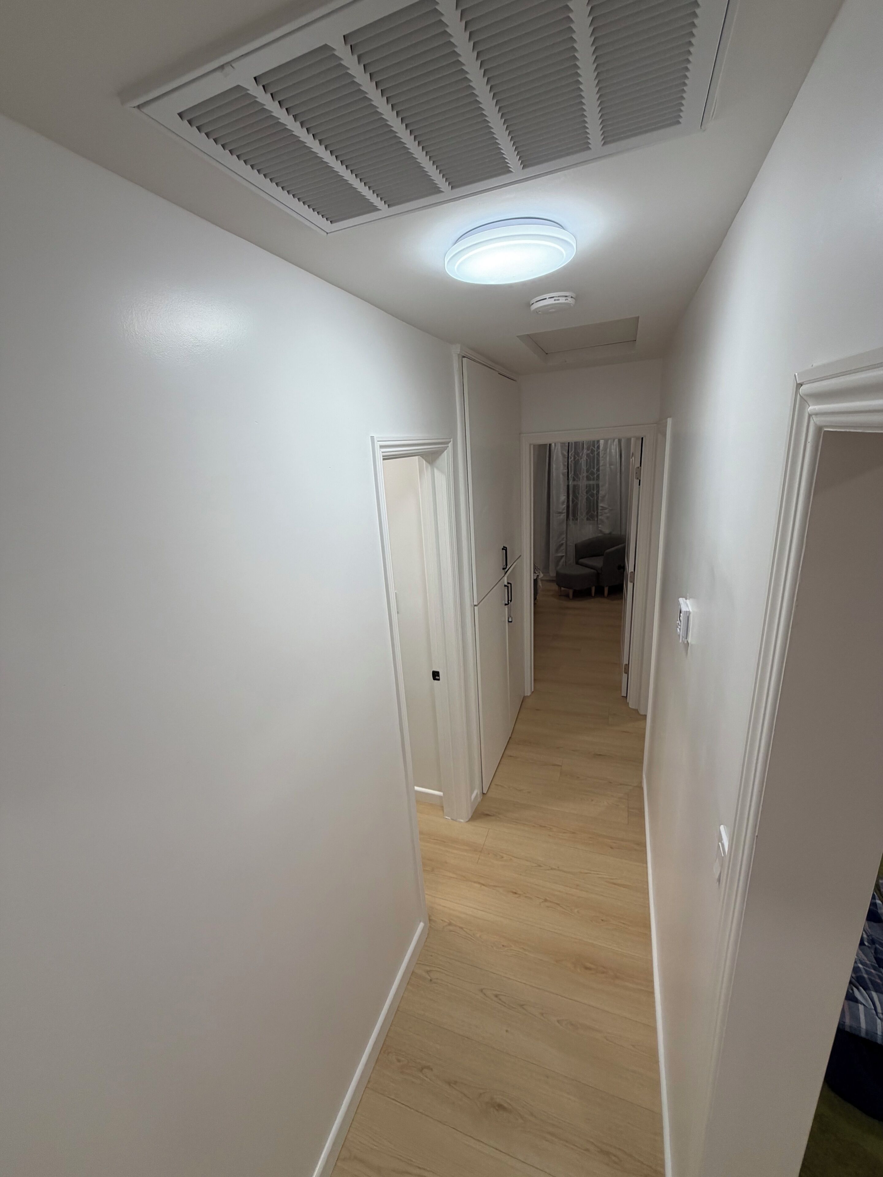 A hallway with white walls, a wooden floor, and a ceiling with a vent and a light fixture.