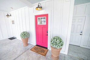 A bright red door with a white panel, flanked by two potted plants with white flowers, and a welcome mat at the entrance. - 6BR, POOL, 2 min walk to beach, Oceanview, Game Rm (Carolina Beach)