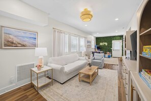 A modern living room with a white sofa, a coffee table, and a TV. There is a green wall and a window with curtains. - 6BR, POOL, 2 min walk to beach, Oceanview, Game Rm (Carolina Beach)