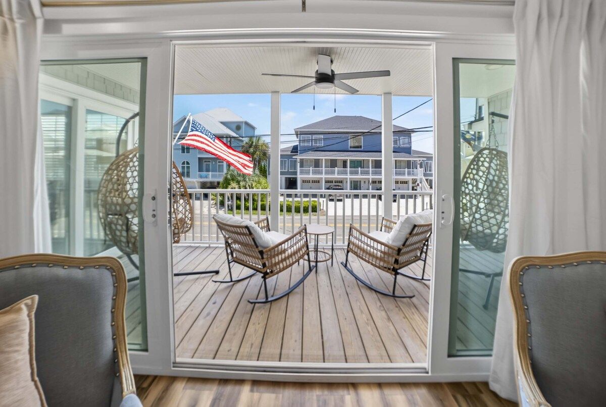 A room with a sliding glass door leading to an outdoor deck with wicker furniture and a view of a house with an American flag.