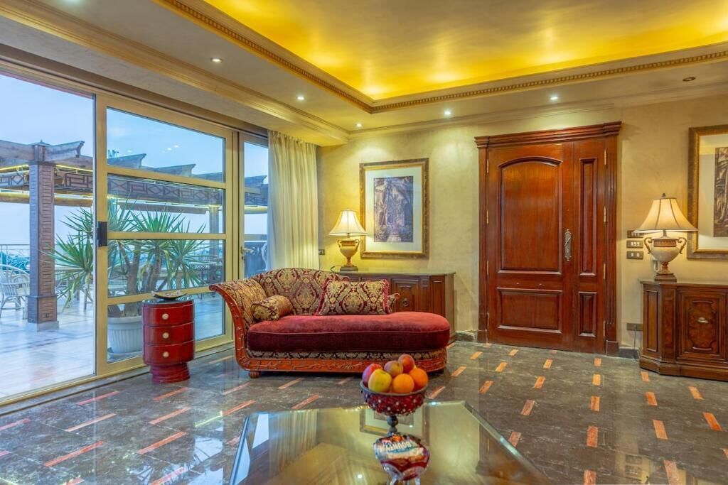 A spacious room with a red sofa, a glass coffee table, and a large window offering a view of palm trees.