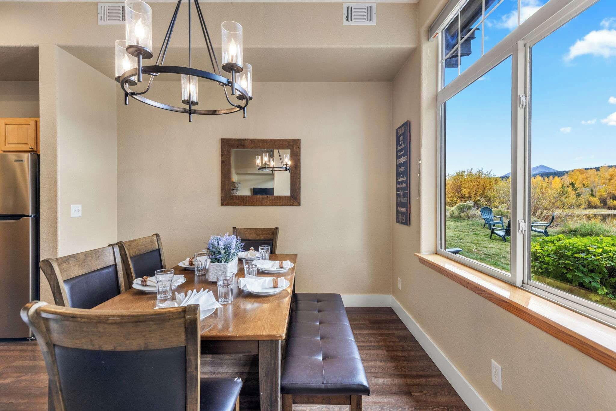 A dining area with a wooden table, chairs, a bench, a chandelier, a mirror, and a window with a view of the outdoors.
