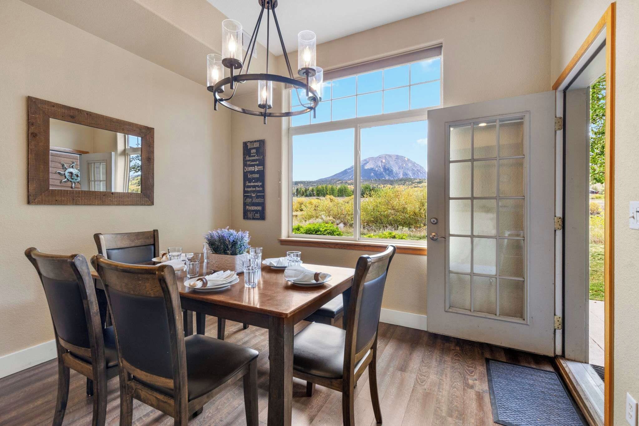 Un coin repas avec une table en bois, des chaises et un lustre. Il y a une fenêtre donnant sur une montagne et une porte avec des panneaux en verre dépoli.