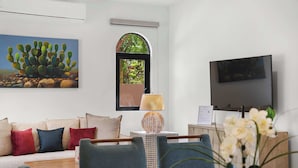 Ein Wohnzimmer mit einer Couch, einem Fernseher, einem Bild einer Kaktee und einem Fenster mit Blick auf Bäume.