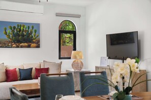 Ein Wohnzimmer mit einer Couch, einem Fernseher, einem Bild einer Kaktee und einem Fenster mit Blick auf Bäume.