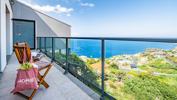 Un balcon avec vue sur la mer et sur la ville. On y trouve deux chaises en bois et un sac rose sur lequel est écrit le mot « HOME ».
