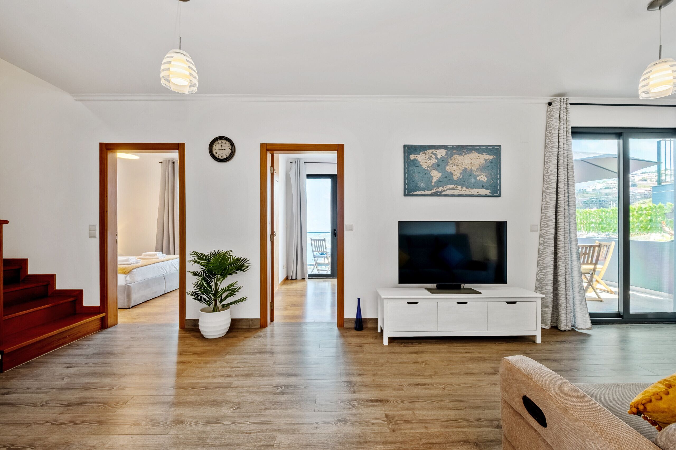 A modern living room with a wooden floor, a television on a white console, a potted plant, and a view of the outdoors through a large window.