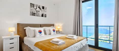 Habitación de hotel con una cama grande, una mesita de noche con lámpara, un cuadro en la pared y un balcón con vistas.