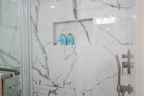 A bathroom with a marble wall, a glass shower enclosure, and a shelf with two bottles of argan oil.