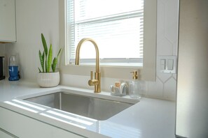 A kitchen with a stainless steel sink, a gold faucet, a potted plant, and a window with blinds.