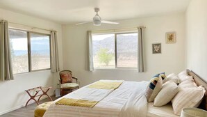 Un dormitorio con cama, una silla, ventilador de techo y dos ventanas con cortinas.