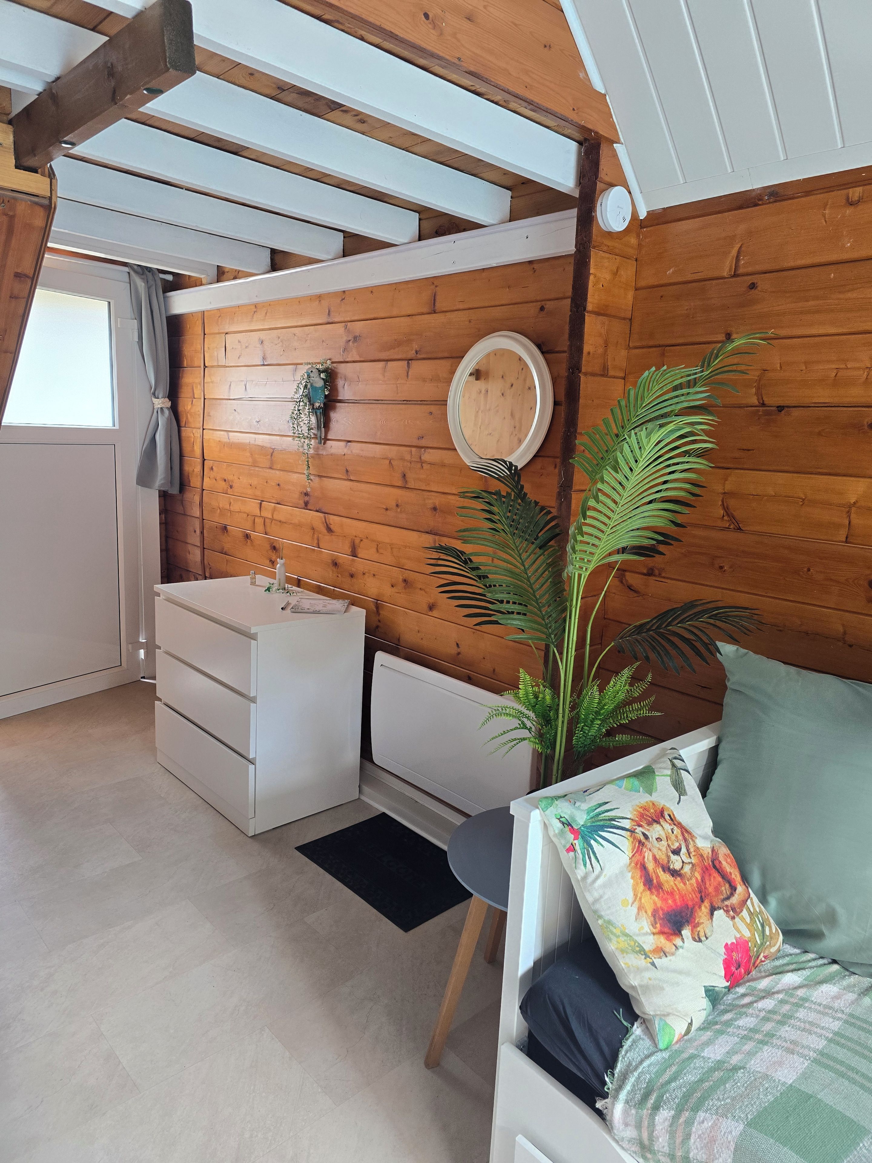 Ein gemütliches Schlafzimmer mit Holzbalkendecke, einem weißen Schminktisch, einem runden Spiegel, einer Topfpflanze und einem Bett mit einem blumigen Kopfkissen.