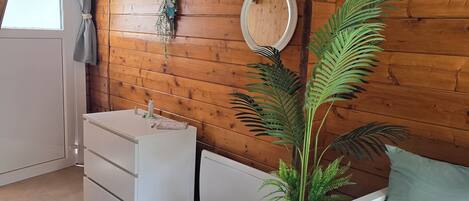 Un dormitorio acogedor con techo de madera, cómoda blanca, espejo redondo, planta en maceta y cama con almohada estampada.