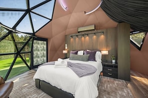 A bedroom with a large skylight, a bed with pillows, a nightstand, and a wall-mounted air conditioning unit. - Evergreen Escape | Private Glass Dome, Sauna & Forest Views (Redmond)