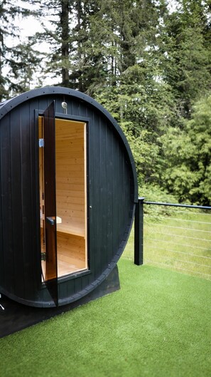 A cylindrical wooden structure with an open door, situated on artificial grass with a fence and trees in the background. - Evergreen Escape | Private Glass Dome, Sauna & Forest Views (Redmond)