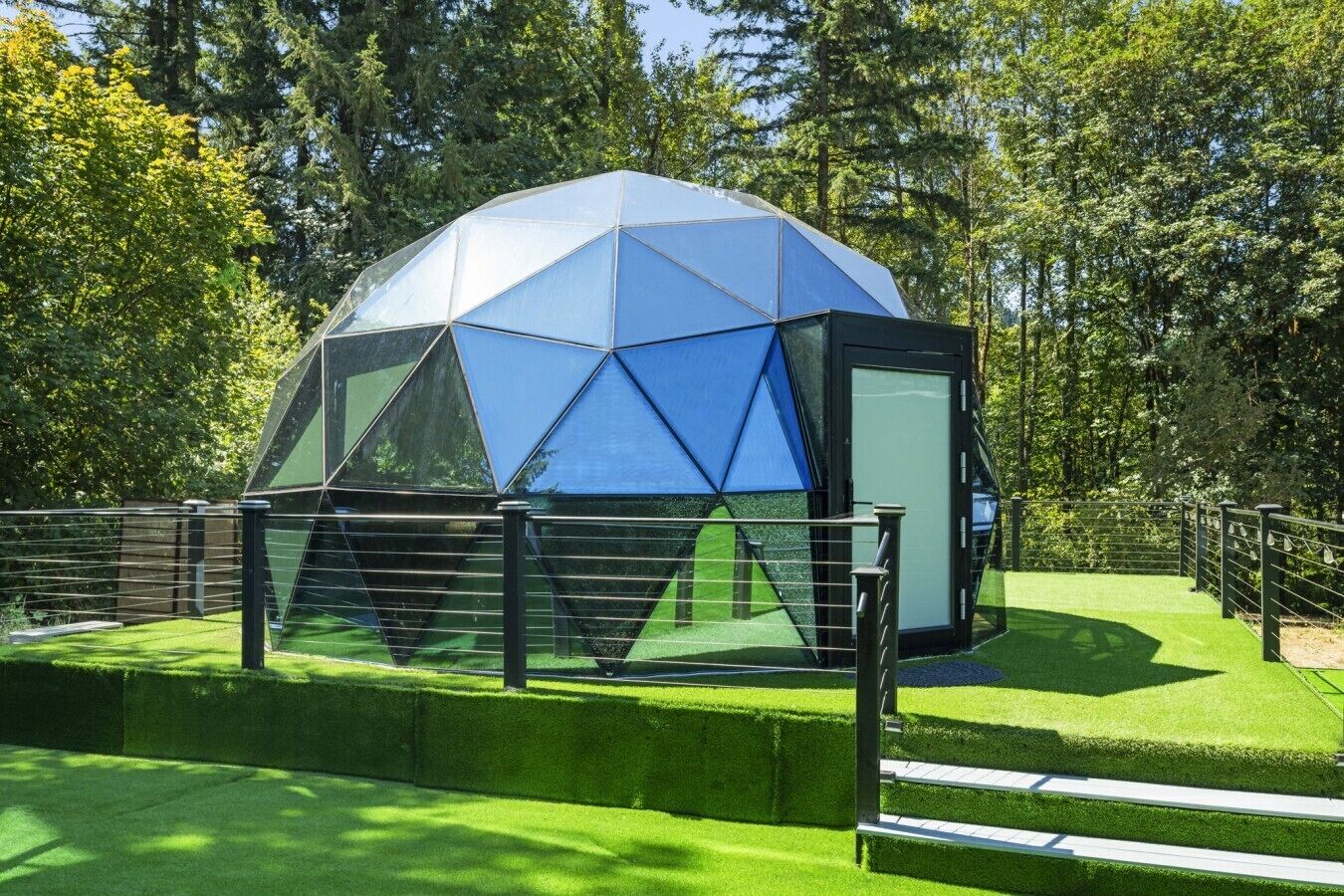 Estructura en forma de domo geodésico con techo transparente, rodeada por una cerca metálica y césped artificial verde.