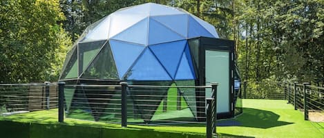 Estructura en forma de domo geodésico con techo transparente, rodeada por una cerca metálica y césped artificial verde.