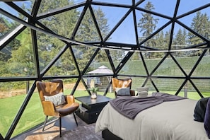 A geodesic dome room with a bed, two chairs, a table, and a view of a green landscape.