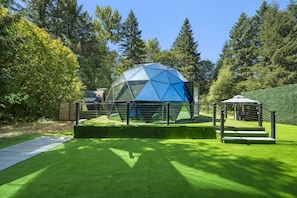 A geodesic dome structure with a surrounding fence and a small pavilion in a landscaped area with grass and trees.