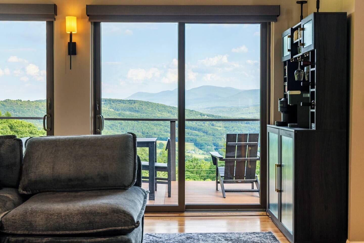 A room with a sofa, a table, and a chair facing a large sliding glass door leading to a balcony with a view of green hills.