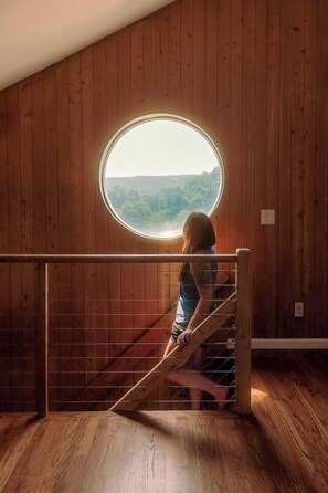 A wooden interior with a circular window overlooking a forested area. - Hollow Sky: 110 Acres, Hot Tub & Views (Rensselaerville)