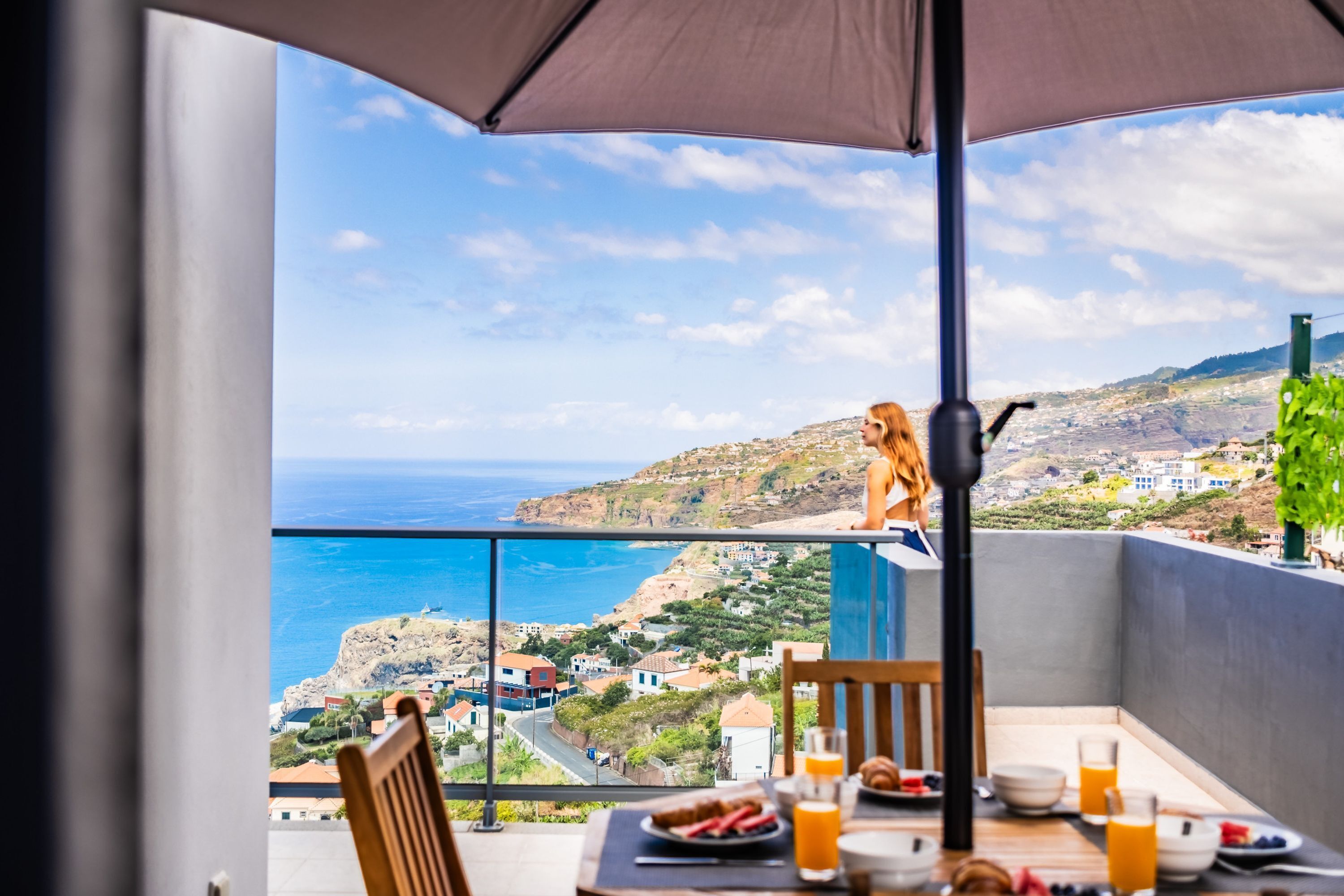 A balcony with a table set for breakfast, overlooking a coastal landscape with houses and cliffs.