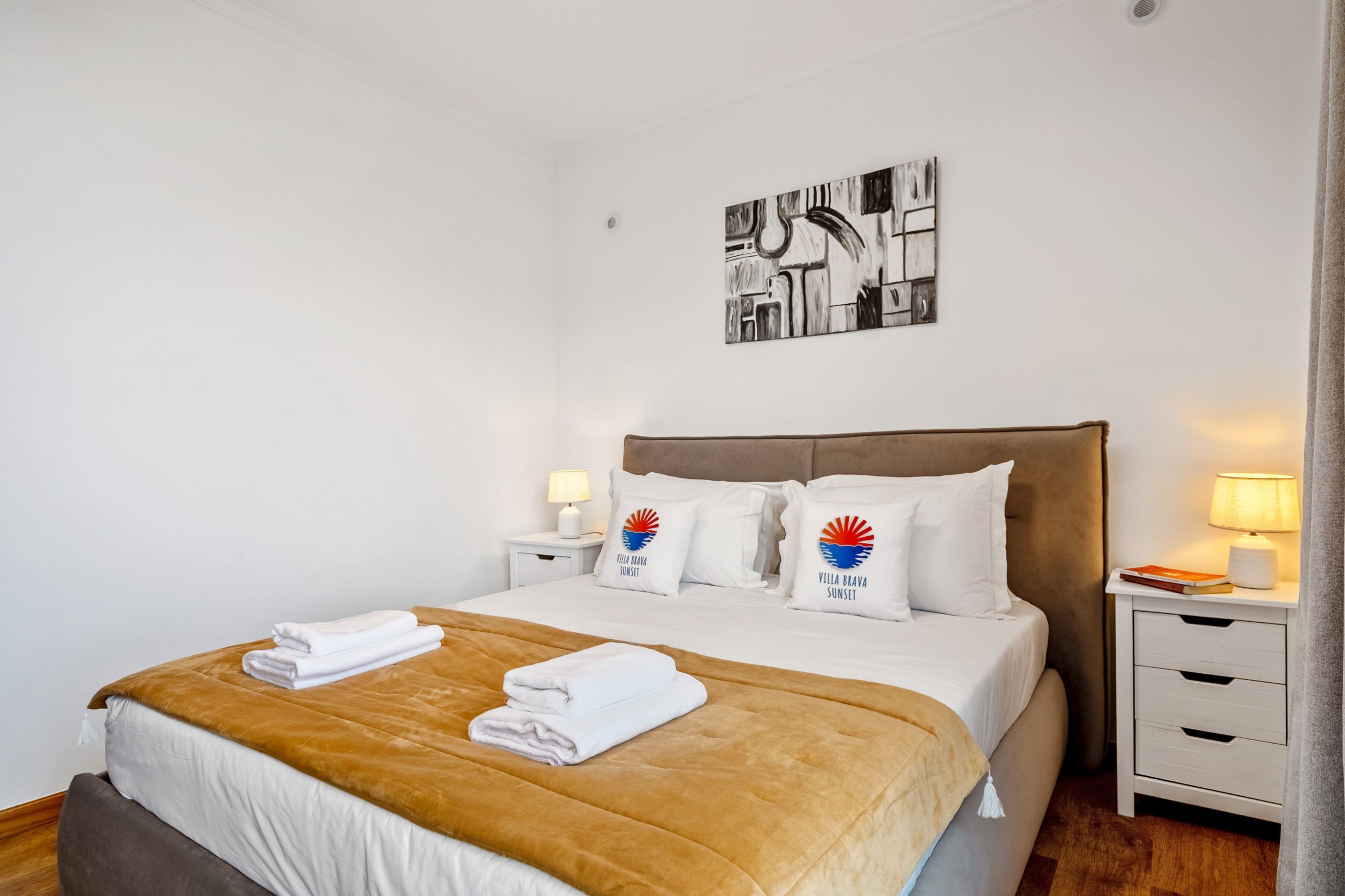 Une chambre d’hôtel comprenant un lit, deux tables de chevet, une tête de lit, une œuvre d’art murale et une fenêtre avec des rideaux.