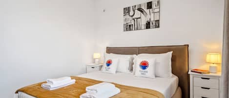 Une chambre d’hôtel comprenant un lit, deux tables de chevet, une tête de lit, une œuvre d’art murale et une fenêtre avec des rideaux.
