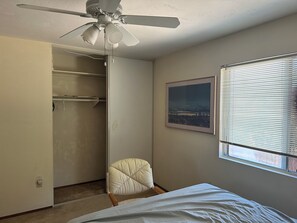Ein Schlafzimmer mit einem Bett, einem Sessel, einem Deckenventilator, einem Fenster mit Jalousien und einem gerahmten Bild an der Wand.