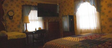 A bedroom with a bed, a desk, a chair, a lamp, a television, and curtains.