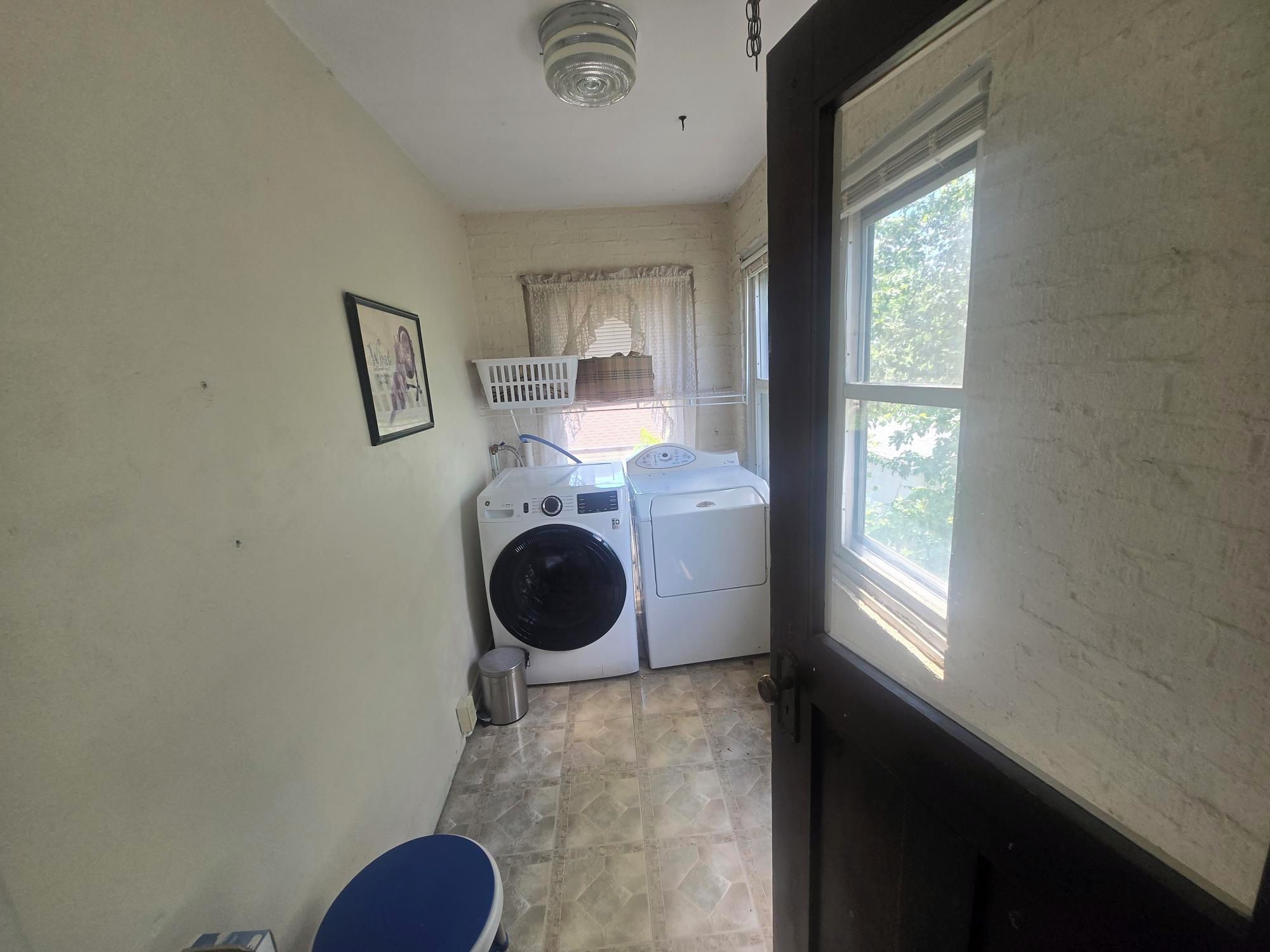 A laundry room with two washing machines, a drying rack, a bucket, and a window with a view of greenery.