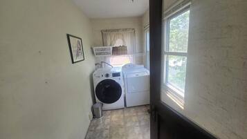 A laundry room with two washing machines, a drying rack, a bucket, and a window with a view of greenery.