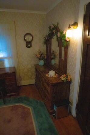 Una habitación con un aparador de madera, un espejo con un arreglo floral, un reloj de pared y una ventana con persianas.