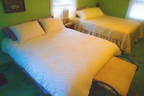 A room with two beds, each with white bedding and pillows. There is a window with curtains, a bedside table with a lamp, and a wall-mounted air conditioning unit.