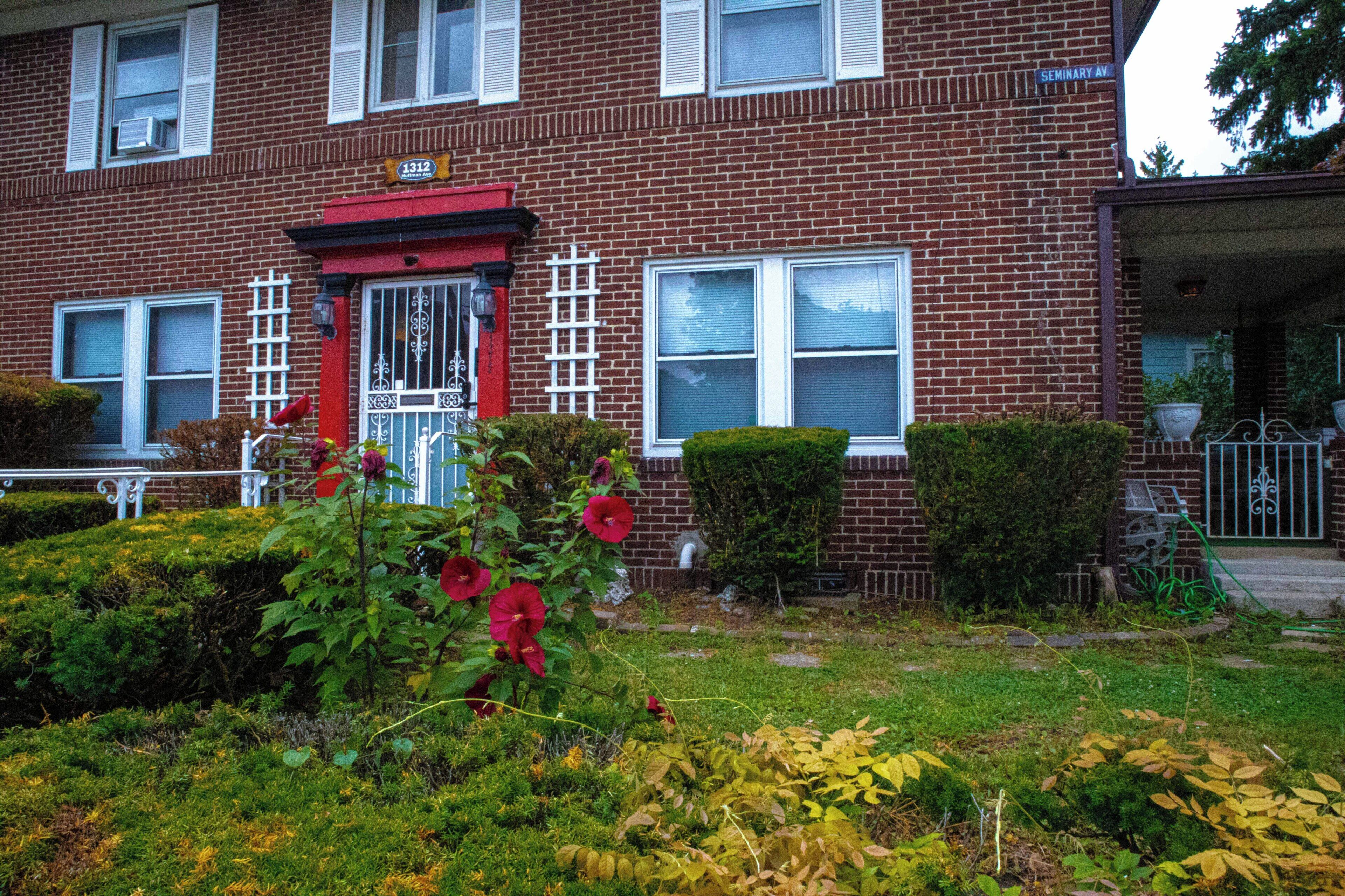 A two-story brick building with a red door and white shutters, a visible address sign, and a well-maintained front yard with various plants and a small garden.