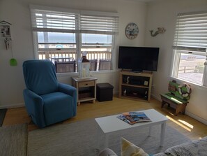 A living room with a blue armchair, a television on a wooden stand, a white coffee table, and a view of the beach through large windows. - OC Md family condo, 19th bdwlk- Sleeps 8, 3 bedroom, 2 bath, Positive reviews. (Ocean City)