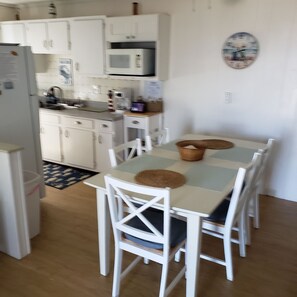 Een keuken met witte kastjes, een magnetron en een koffiezetapparaat. Er is een eettafel met witte stoelen en een geweven placemat.