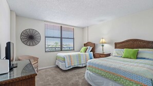 A bedroom with two beds, a television, a lamp, and a wicker chair. - Sandy Toes l Oceanfront Condo l Heated Pool (St. Augustine)